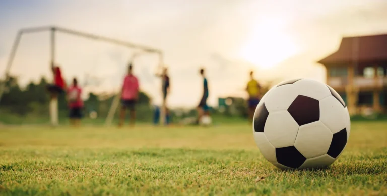 A soccer ball sits on a grassy field with a goalpost and people blurred in the background during sunset.