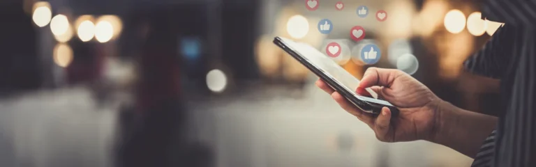 Close-up of a person holding a smartphone with floating Facebook-like thumbs-up and heart icons on a blurred background.