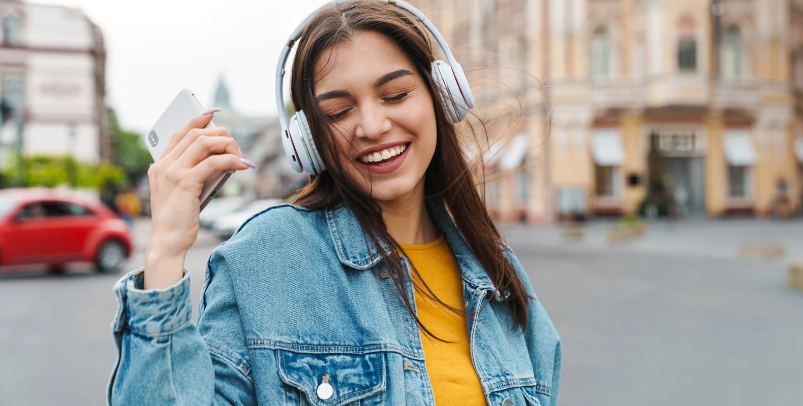 Woman outdoors wearing headphones, holding a smartphone, smiling, and dancing with her eyes closed.