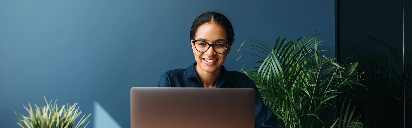 Smiling person wearing glasses, seated at a desk, using a laptop, with green plants in the background.