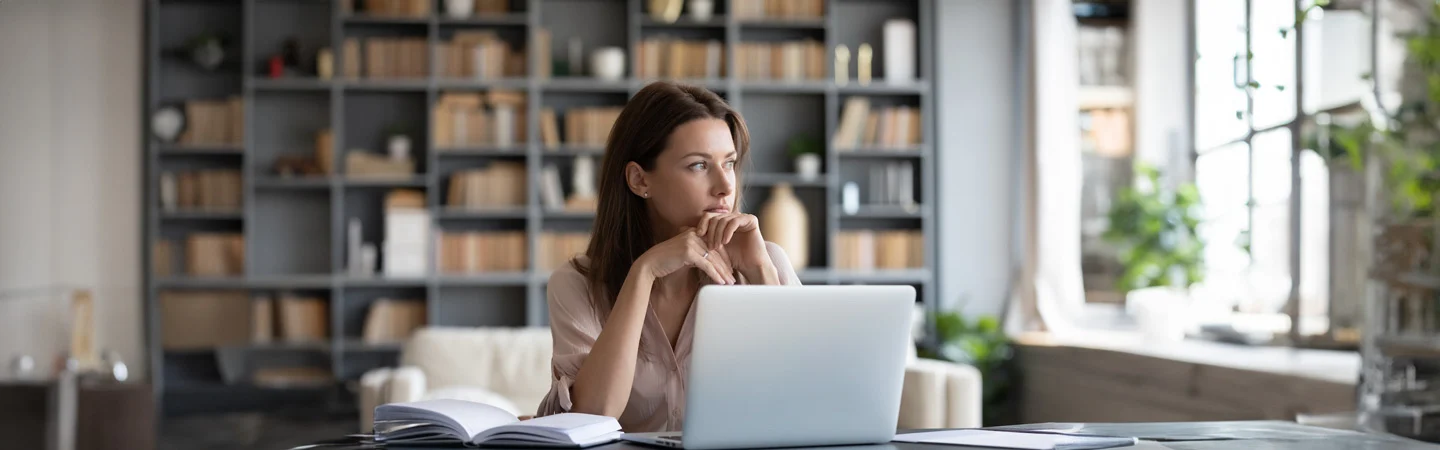 A woman sitting at a desk in a library, looking thoughtful with an open book and a laptop in front of her.