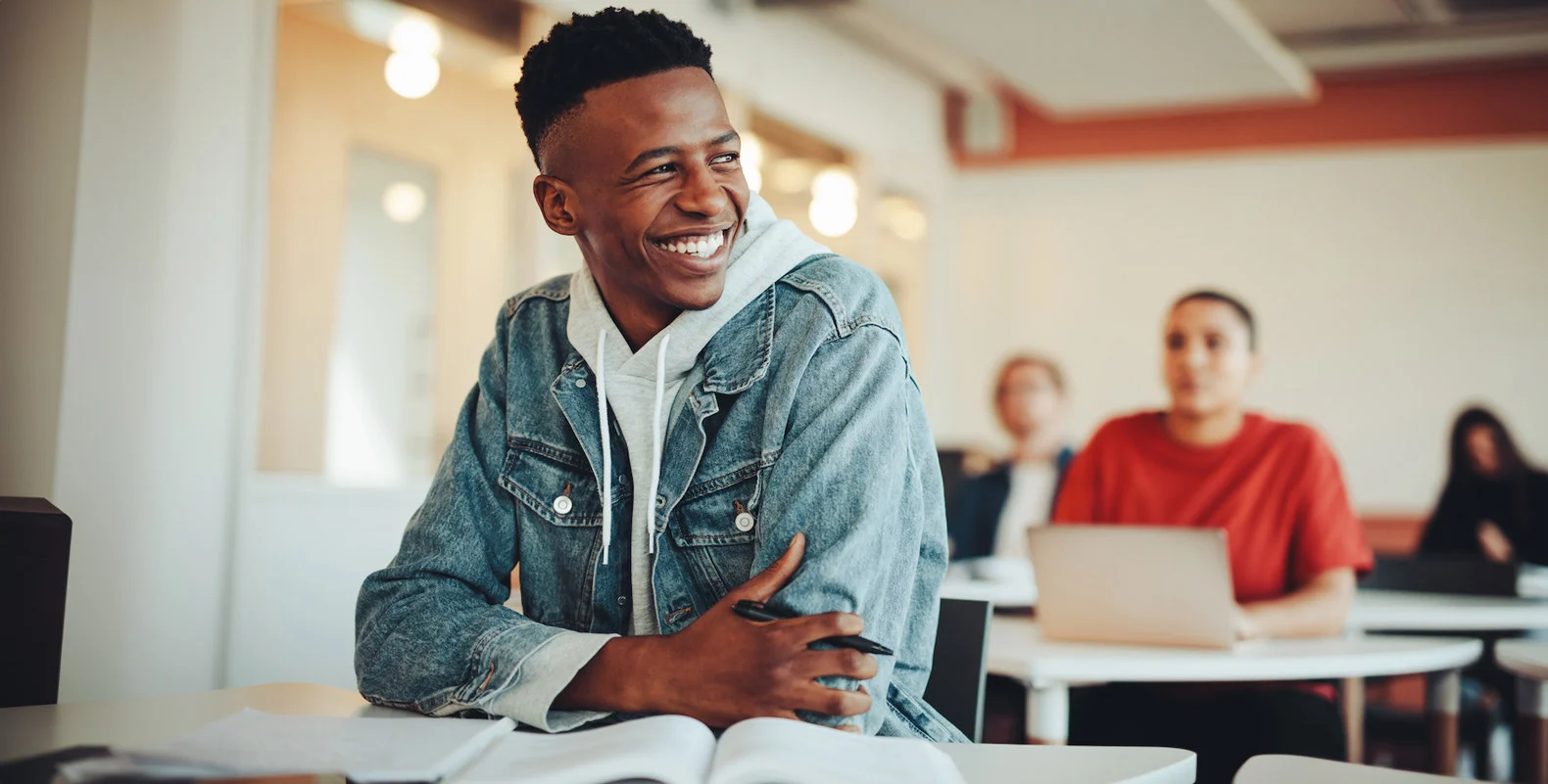 Smiling student in a denim jacket sits in a classroom. Others are seated in the background, with one working on a laptop.