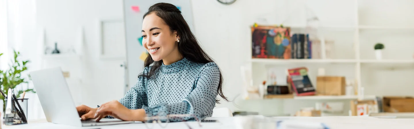 Woman in a polka dot blouse smiling, working on a laptop at a desk in a bright, organized office space.