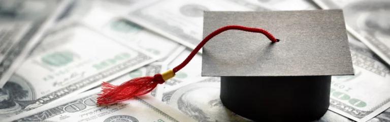 A graduation cap with a red tassel on top of scattered 100-dollar bills.