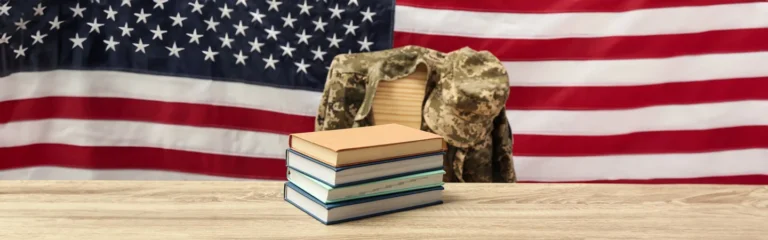 Books stacked on a table with a military uniform jacket draped over a chair and an American flag in the background.