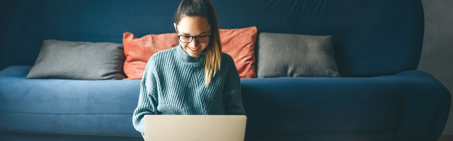 Person smiling and working on a laptop while sitting on the floor in front of a couch with colorful pillows.