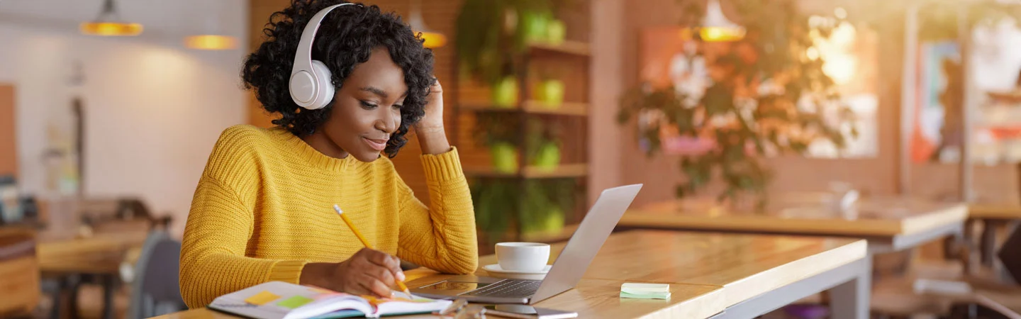 A person wearing headphones and a yellow sweater studies with a laptop at a well-lit café.