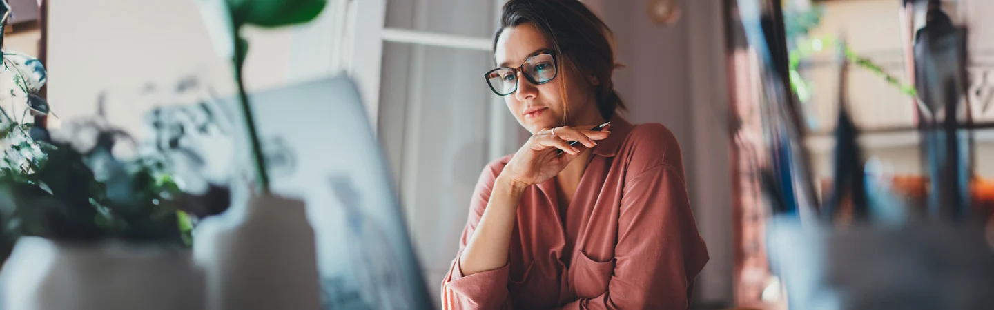 A woman in glasses and a pink shirt reflects thoughtfully while looking at a laptop in a cozy, plant-filled room.