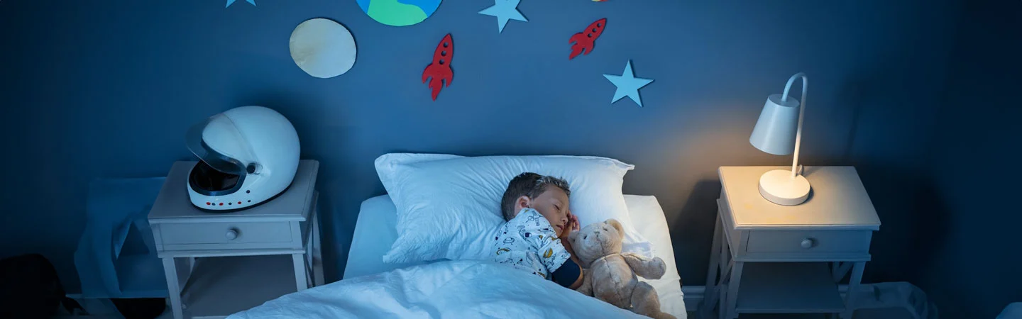 Child sleeps in bed holding a teddy bear, surrounded by space-themed decorations and a helmet on the bedside table.