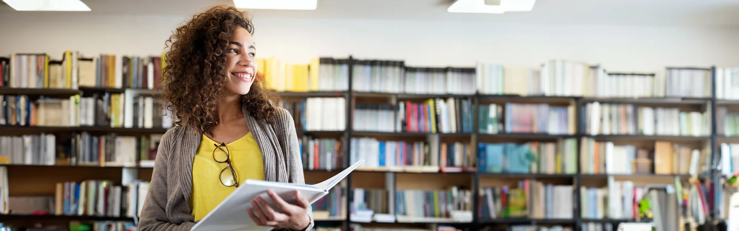 A person with curly hair stands smiling and holding a book in a library, with shelves of books in the background.