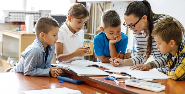 A teacher helps five young students with their work at a table in a classroom.