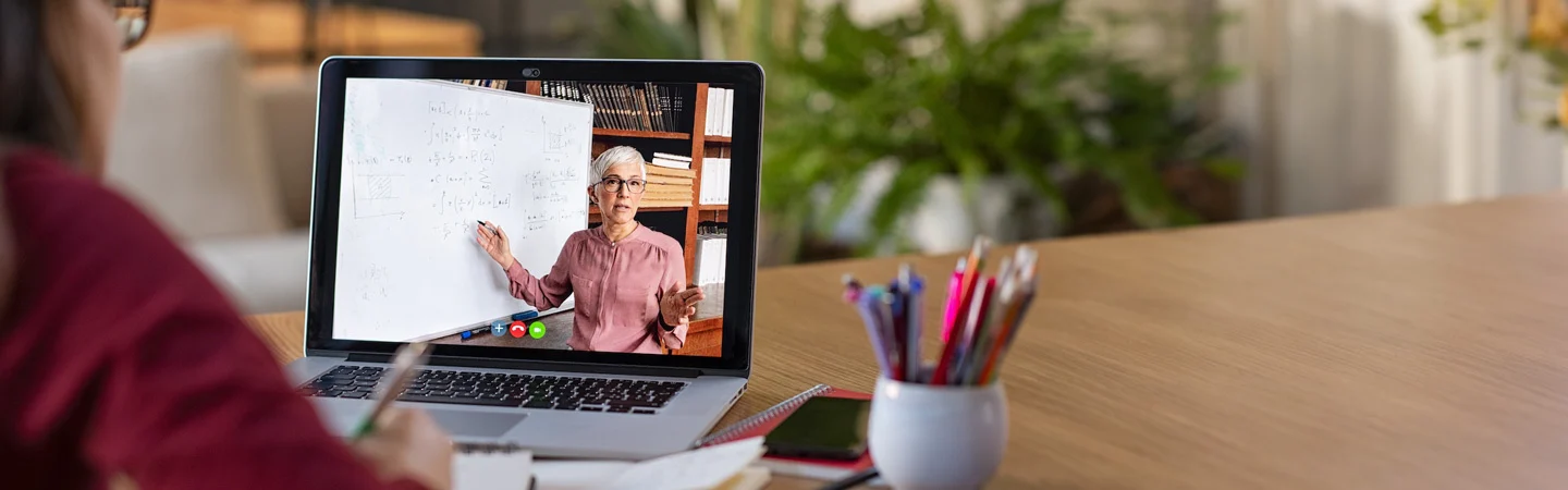 Person watching an online class on a laptop in a home setting, with a whiteboard and teacher visible on screen.