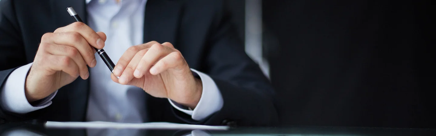 A person in a suit holding a pen, sitting at a desk with papers, against a dark background.