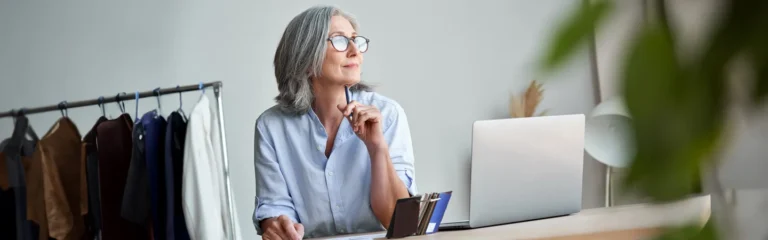 Gray-haired woman in glasses, sitting at a desk with a laptop, holding a pen, and looking thoughtfully to the side.