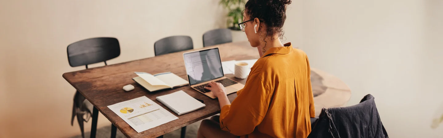 A woman in a mustard shirt works on a laptop at a wooden table with documents, a notebook, and a coffee cup.