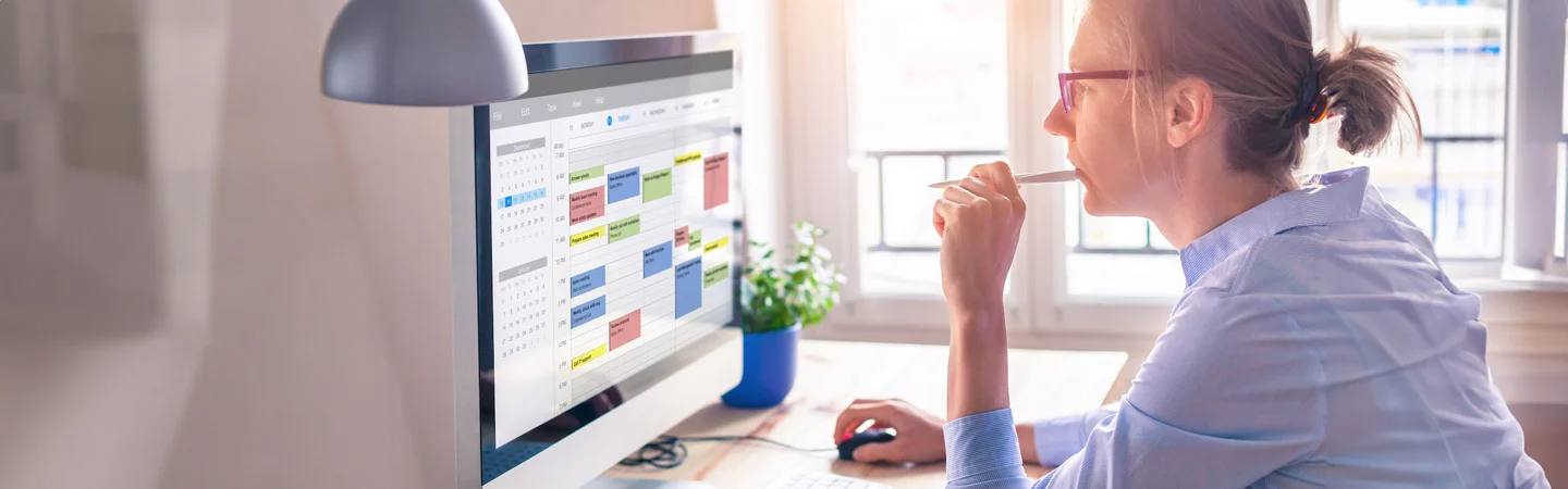 Person working at a desk, looking at a large monitor displaying a calendar with various color-coded events.