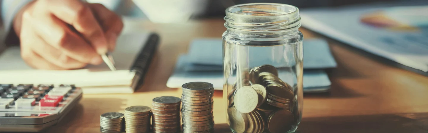 A person is writing while a calculator and stacks of coins leading to a jar of coins are on the desk.