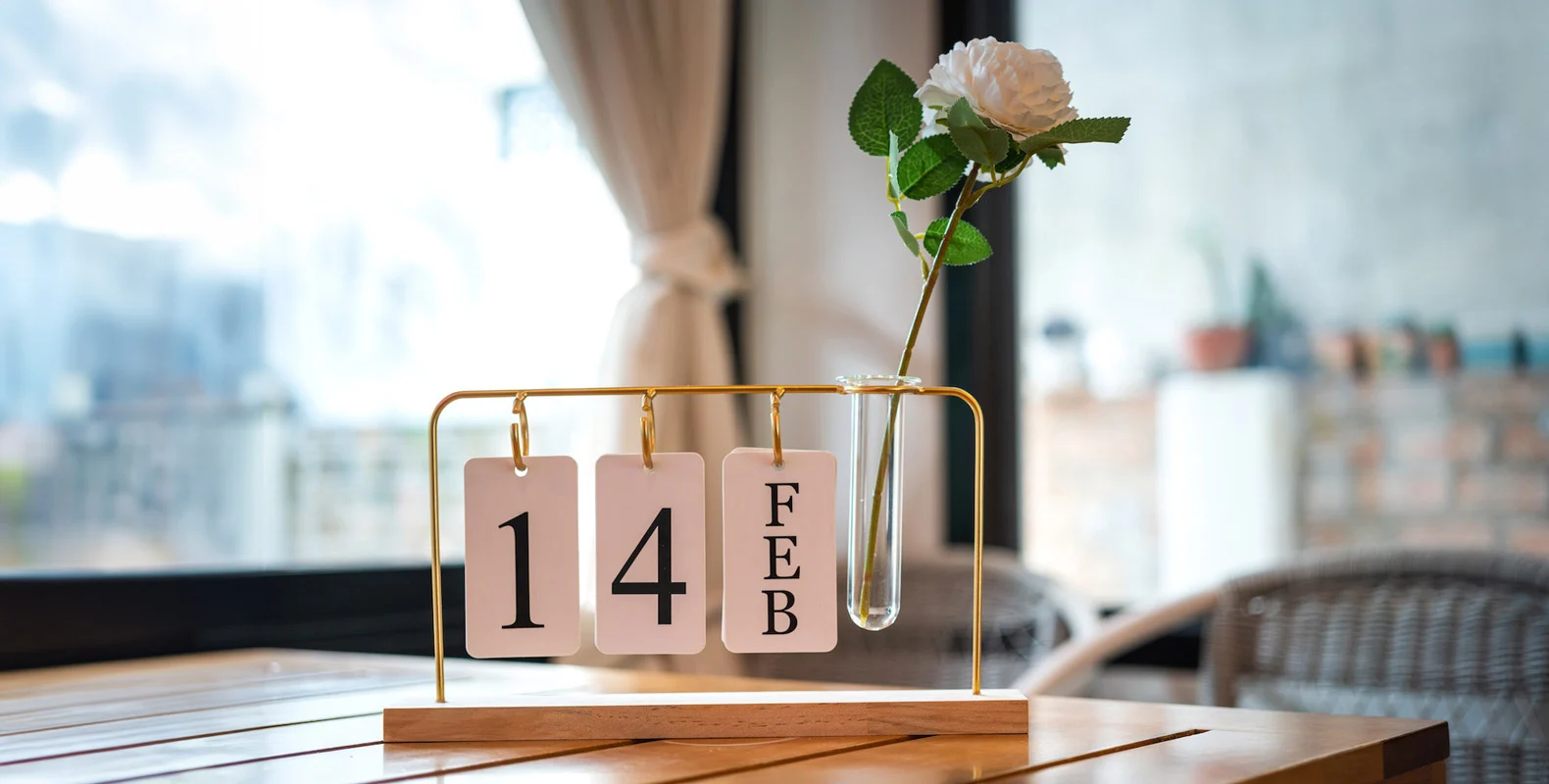 A decorative calendar on a table shows February 14 with a white rose in a vase beside it.