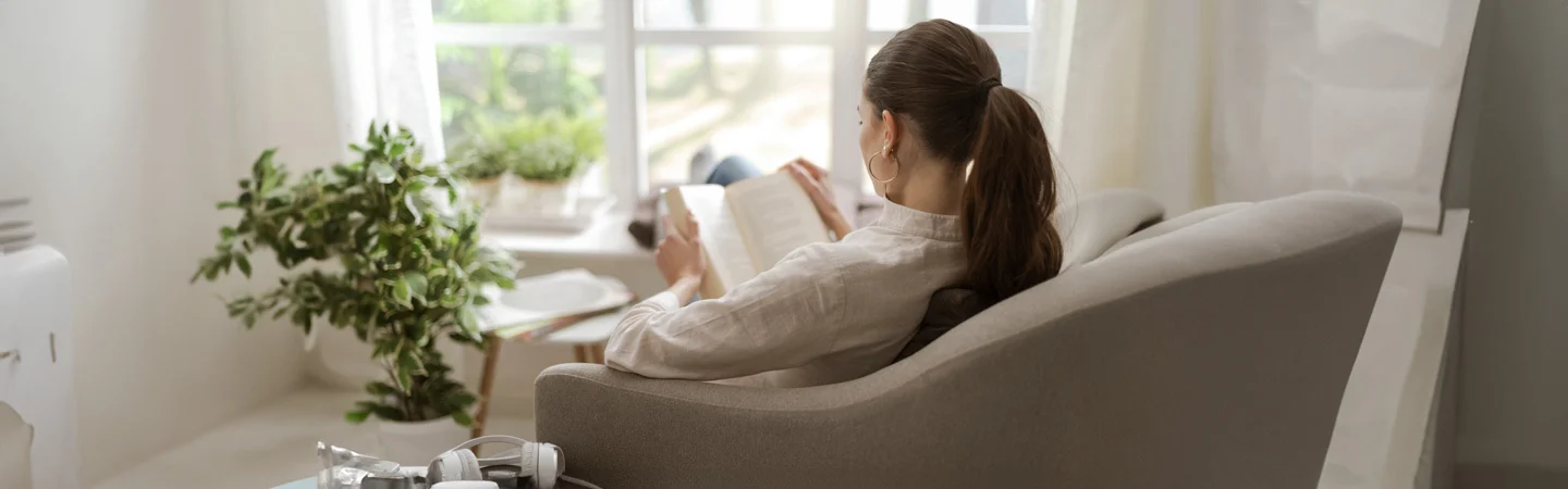 Woman reading a book while sitting in a comfy chair by the window, with a green plant nearby.