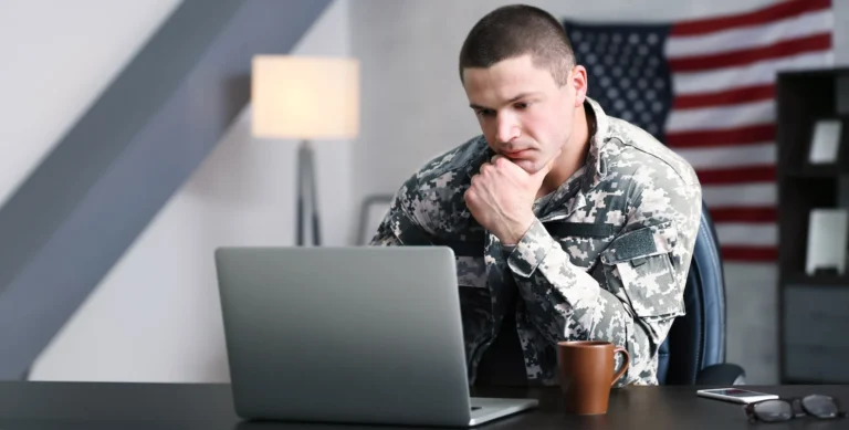 A person in a military uniform is sitting at a desk, looking at a laptop screen with a thoughtful expression. An American flag hangs behind.