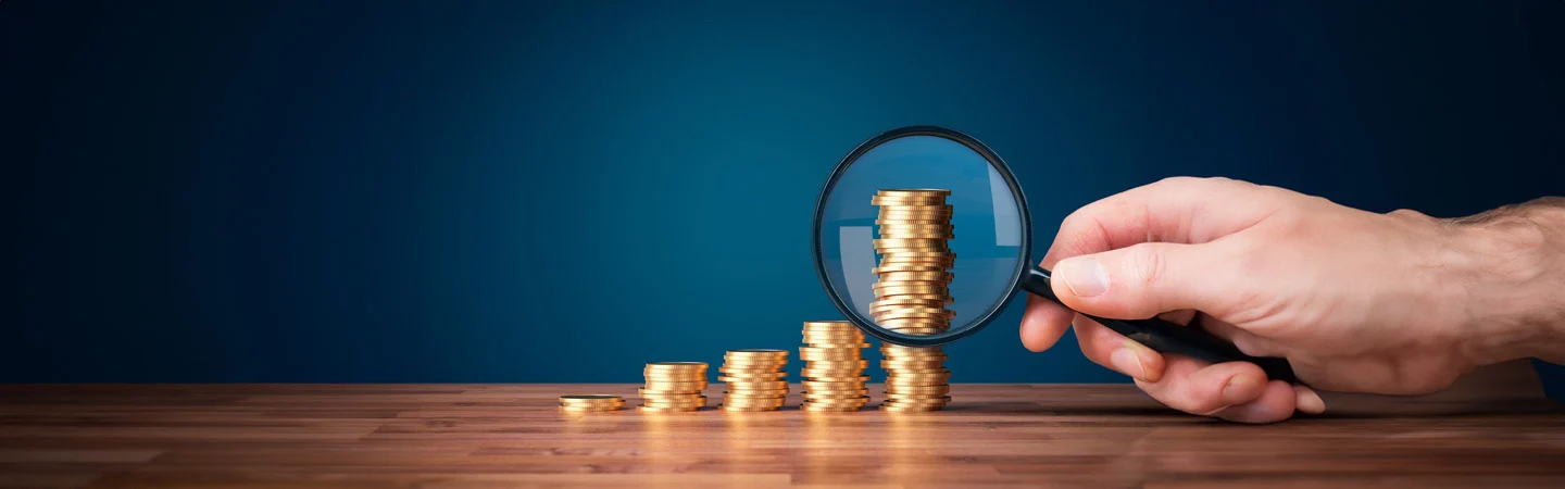 Hand holding magnifying glass over stacked gold coins, symbolizing financial growth and close examination, on wooden surface.