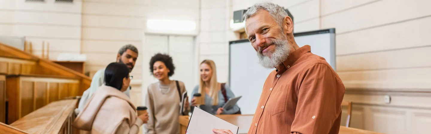 An older man with a tablet smiles in the foreground, while a group of people chat in the background in a bright room.