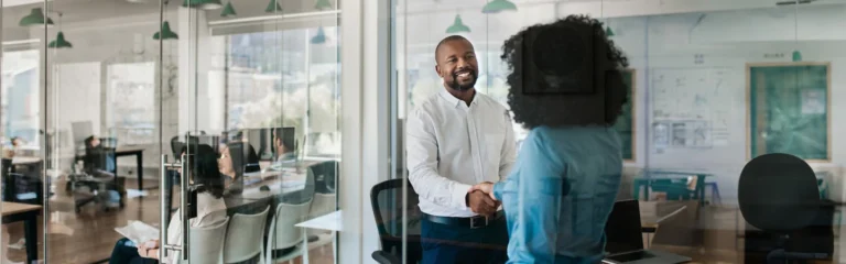 Two people shaking hands in a modern office with coworkers in the background working at their desks.