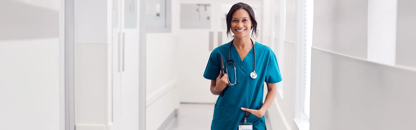 Smiling medical professional in scrubs holding a stethoscope, standing in a bright hospital hallway.