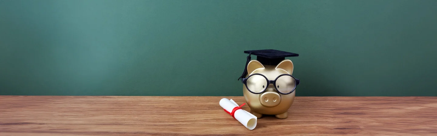 Piggy bank wearing glasses and a graduation cap beside a diploma on a wooden surface against a green background.