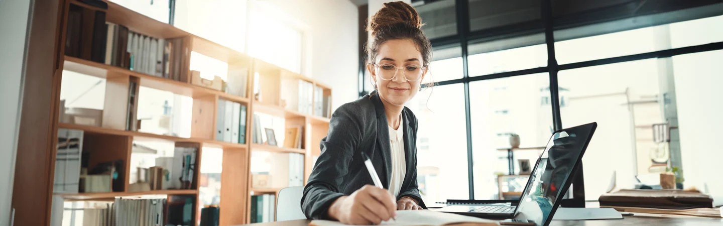 A woman in glasses writes in a notebook at a desk with a laptop in a well-lit office.