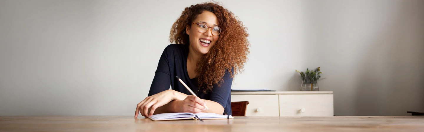 Woman with curly hair and glasses sits at a table, smiling and writing in a notebook.