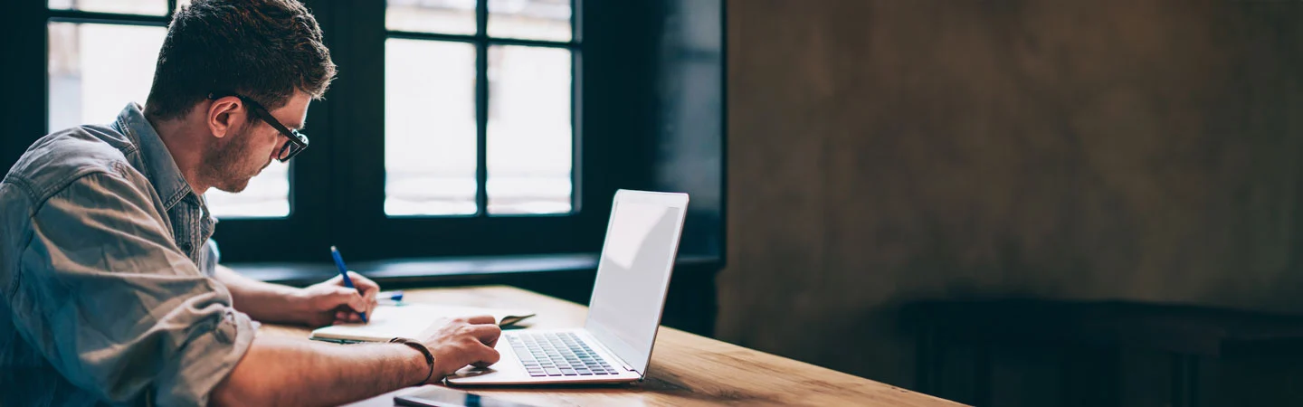 A person is sitting at a wooden table, working on a laptop and taking notes with a pen in hand, near a window.