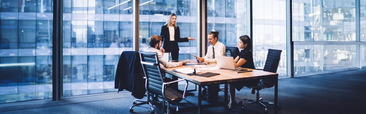 Business meeting with four people in a modern office with large windows, with one person standing and speaking.