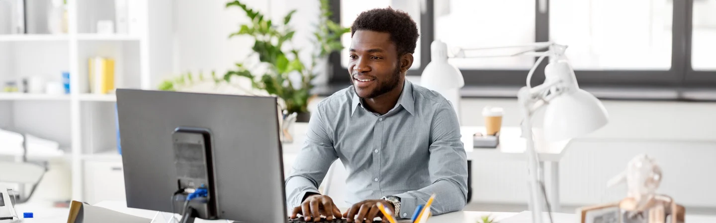 Man smiling while working on a computer in a modern, bright office space with plants in the background.