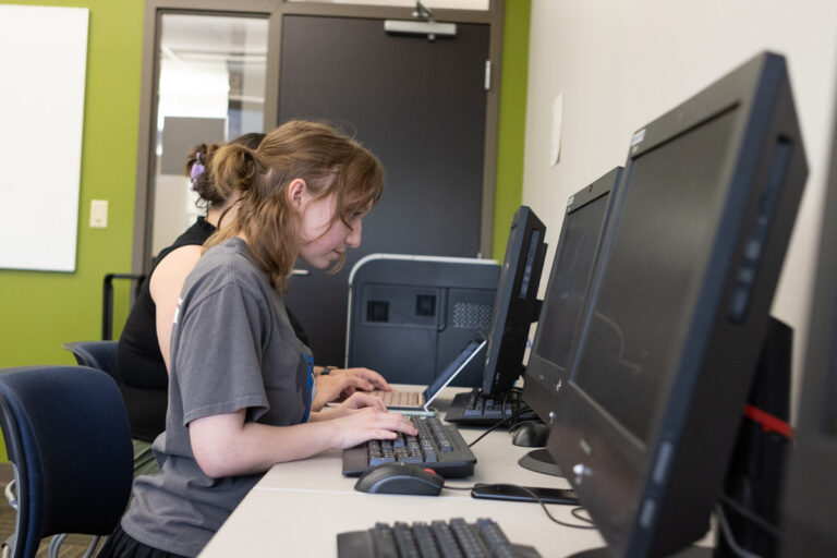 Two people sitting side by side, working on computers in a room with green and white walls and a closed door behind them.