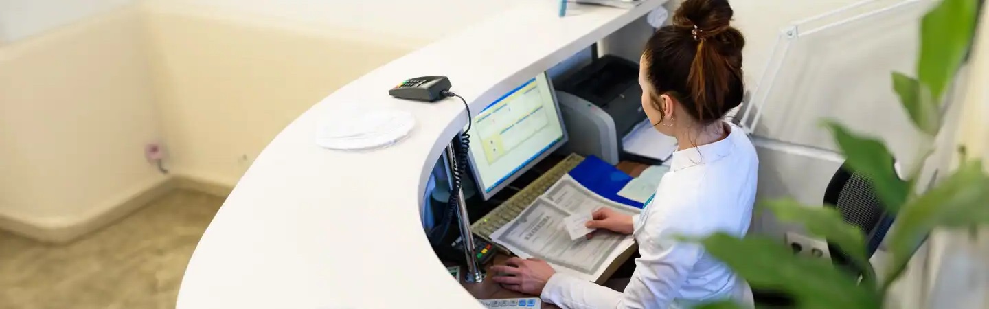 Receptionist working at a curved desk, using a computer while holding a document.