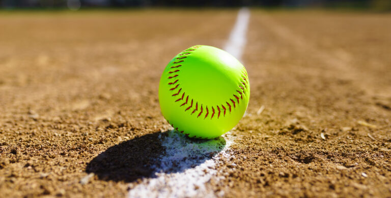 Bright yellow softball lying on a dirt field with a white chalk line underneath it.