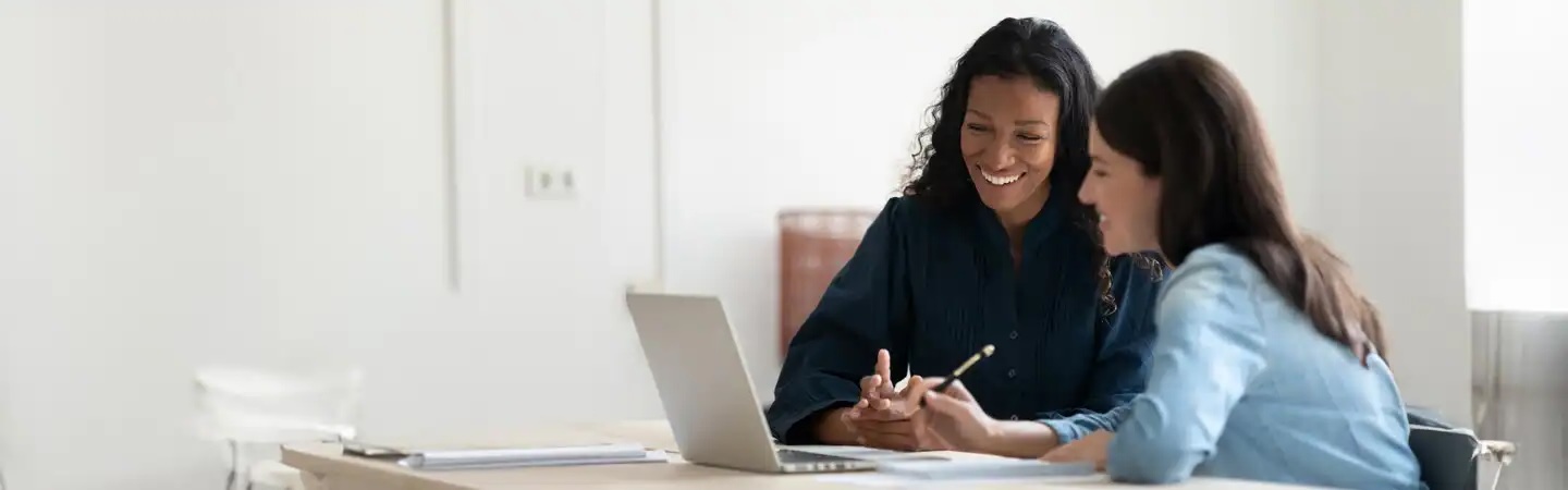 Two women sitting at a table with a laptop, smiling and discussing paperwork in a brightly lit office setting.