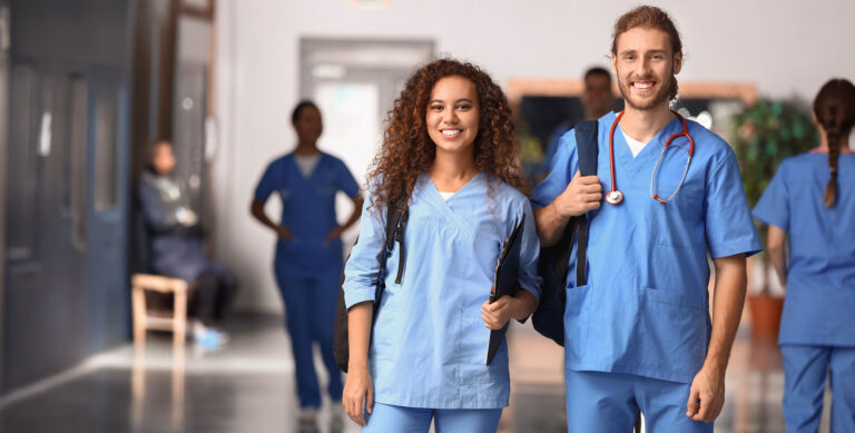 Two smiling nursing students in blue scrubs stand together in a hospital hallway, ready for their shift.