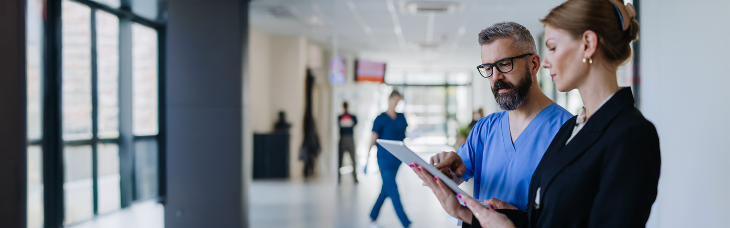 A doctor in scrubs and a woman in formal attire discuss something on a tablet in a hospital corridor.