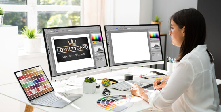 A designer at a desk working on a loyalty card design across two monitors, comparing the creative aspects of digital marketing and graphic design careers.