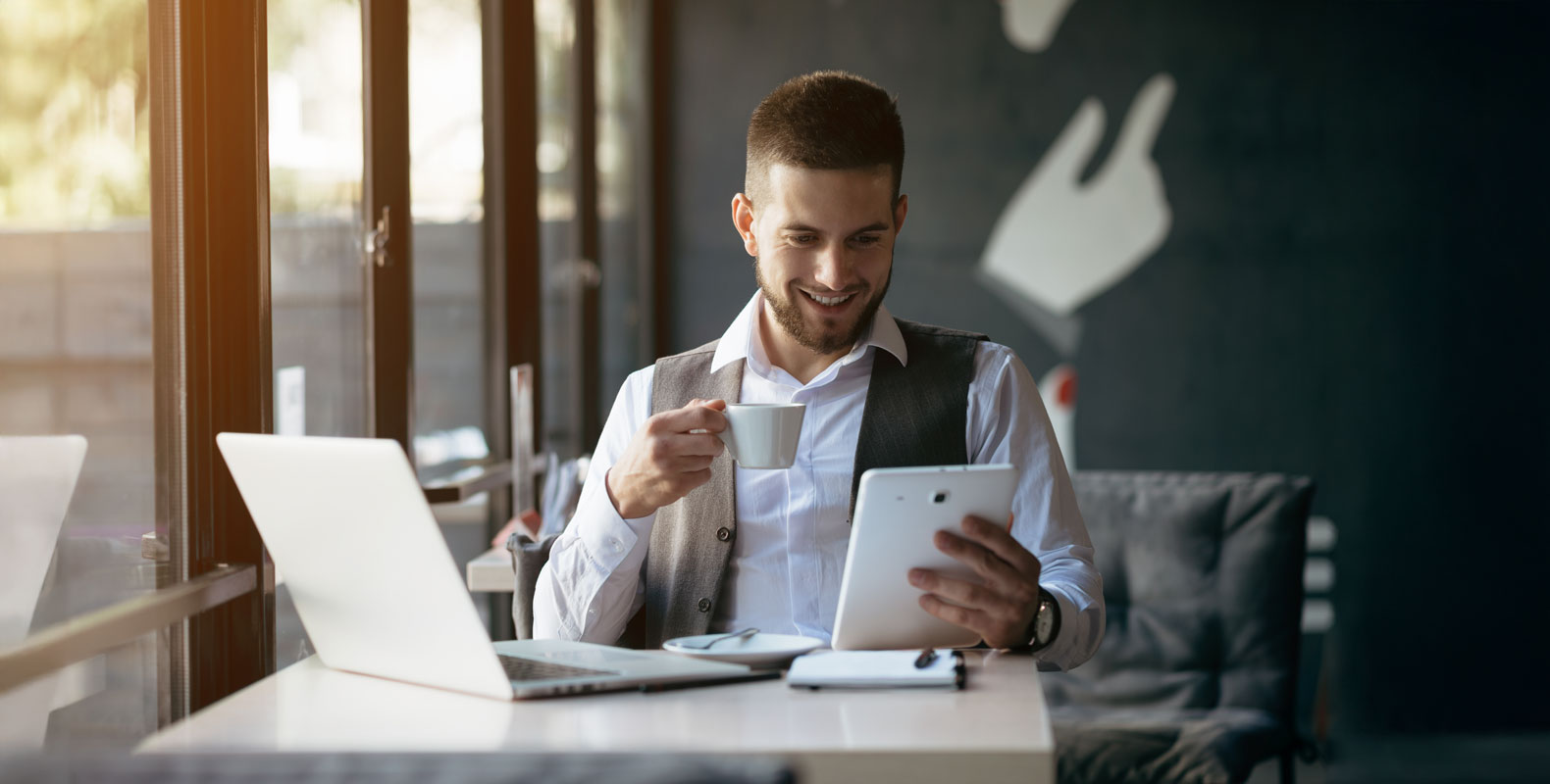 A businessman holding a cup of coffee and working on a tablet in a modern café, representing the flexibility of freelance digital marketing.