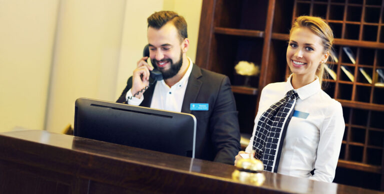 Two hotel front desk employees, one answering the phone and the other smiling at the camera, illustrating key skills learned in hospitality management.