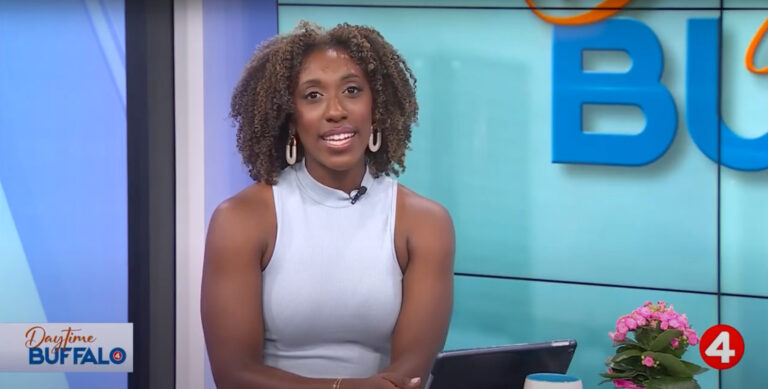 A television presenter smiling during a segment on 'Daytime Buffalo,' with a flower and tablet visible on the desk.