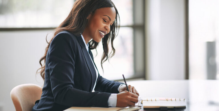 A professional woman in a business suit writing in a notebook at her desk.