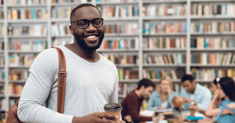 Colleges student in library holding coffee and books with more students and books in the background