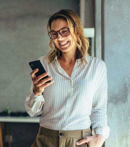 A woman in a white striped blouse and glasses smiles while looking at her smartphone. She is leaning against a gray wall in a well-lit room. Her light brown hair is loose around her shoulders.