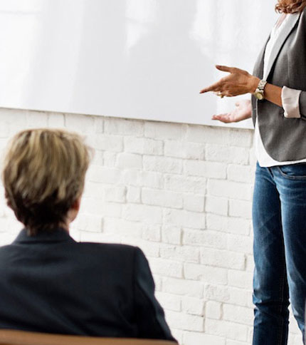 A person in casual business attire stands and gestures while presenting, as another person with short hair sits and listens. The setting appears informal against a white brick wall backdrop.