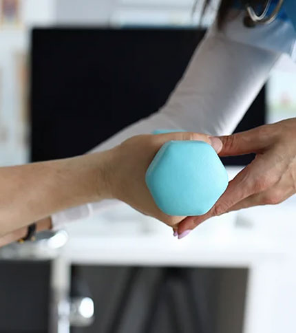 A healthcare professional assists a person with an exercise involving a small blue hexagonal dumbbell. The scene appears to be in a medical or rehabilitation setting, with a focus on physical therapy or rehabilitation.