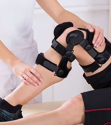 A person wearing a knee brace sits on a medical examination table. Another person, possibly a therapist or doctor, is adjusting the brace. The setting appears to be a clinical or rehabilitation environment.